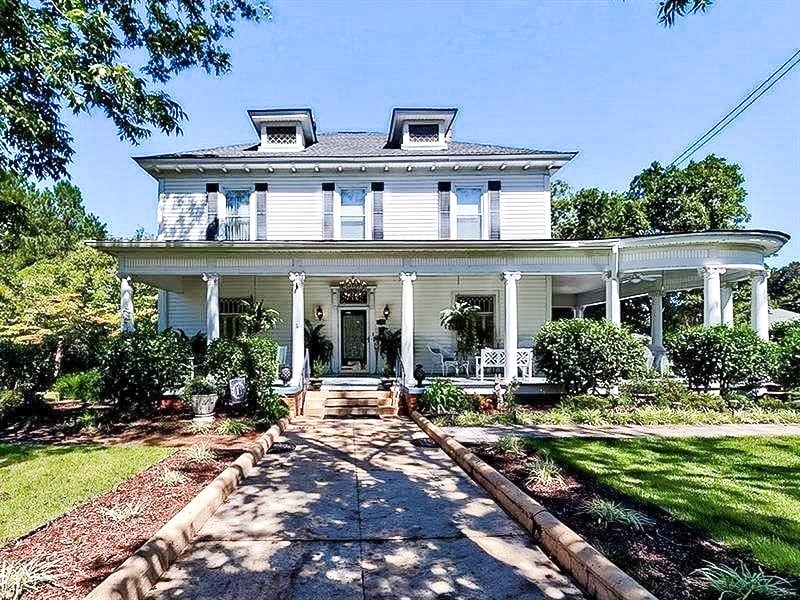 A charming historic home is featured with a large, inviting front porch supported by white columns. Lush greenery surrounds the house, and well-maintained landscaping enhances the appeal. The exterior is finished in light colors, contributing to a welcoming atmosphere.