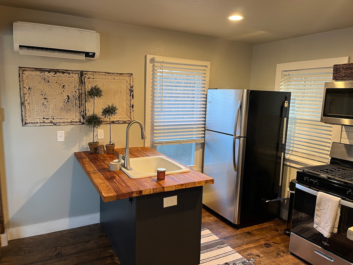 A modern kitchen features an island with a wooden countertop and a sink. Stainless steel appliances, including a refrigerator and stove, are visible. Natural light filters through the window, which is adorned with white blinds. Decorative plants sit on the countertop, adding a touch of greenery.