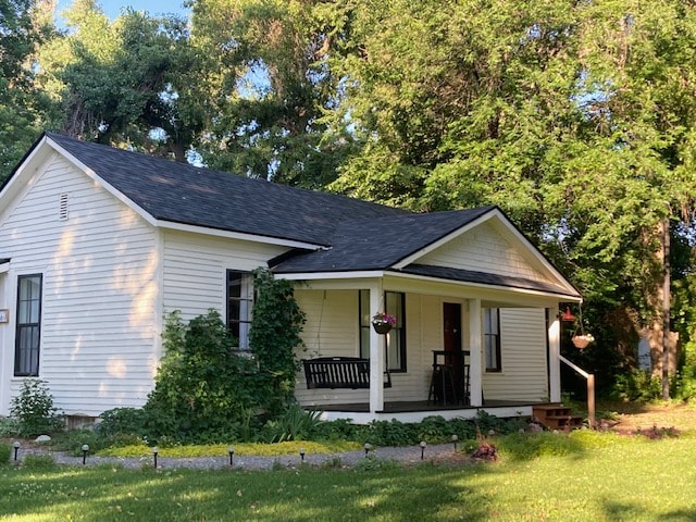 A historic farmhouse is depicted, showcasing a white exterior and a welcoming front porch adorned with hanging flowers. A swing is located on the porch, surrounded by lush greenery. The property is set within a landscaped area, enhancing the natural setting.