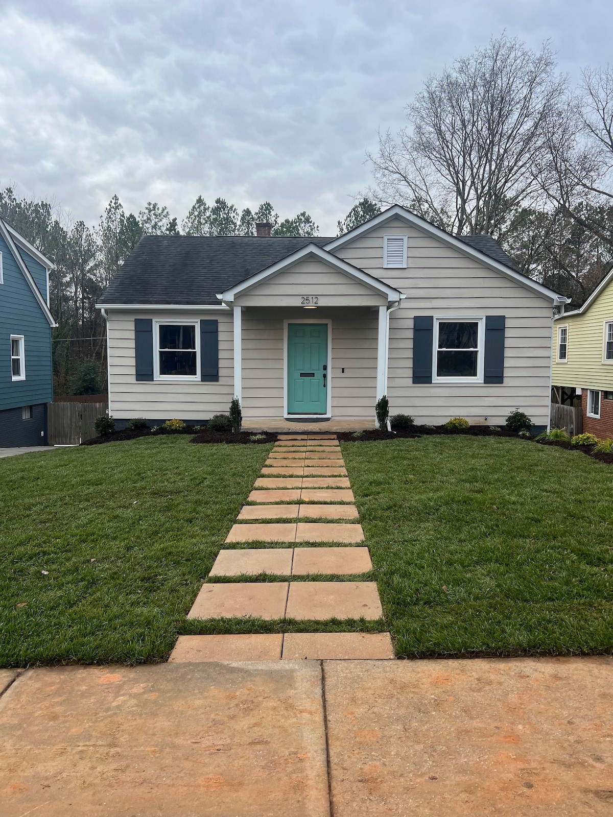A charming bungalow features a light-colored exterior with blue shutters and a mint green front door. A clear stone pathway leads to the entrance, bordered by a well-manicured lawn and planted greenery, creating an inviting front entryway.
