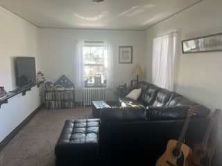 A cozy living area is furnished with a dark leather sectional sofa, complemented by a light-colored rug. Natural light filters through two white-curtained windows, illuminating the space. A small table sits in front of the seating, and a shelf displays various books and decor. Two acoustic guitars rest nearby.