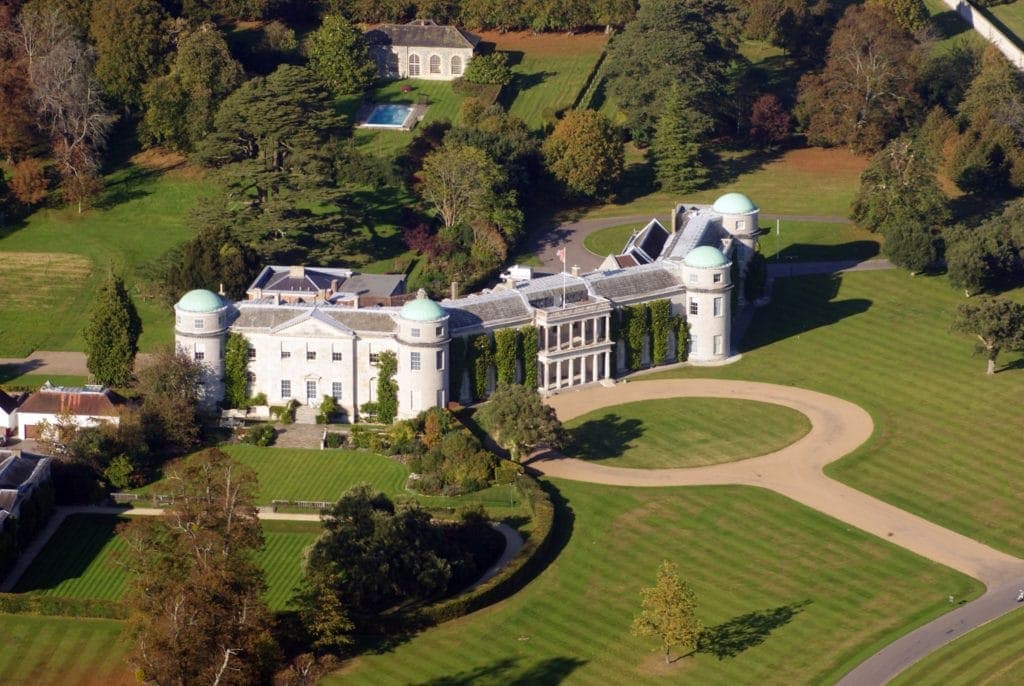 An elegant manor is showcased, surrounded by lush greenery and well-maintained gardens. The symmetrical structure is highlighted by distinctive dome-topped towers, with vast lawns creating a grand entrance. A pool and gazebo can be seen in the landscaped grounds, adding to the estate's charm.