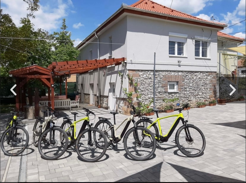 A paved area is visible in front of a charming building, featuring six parked e-bikes in varying colors, including white and bright yellow. Sunlight filters through nearby trees, casting gentle shadows on the ground. A covered seating area with wooden beams is located to the left.