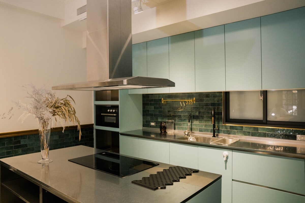 The kitchen features soft blue cabinetry, complemented by a sleek metallic countertop. An island with an electric stovetop is centrally located, while the wall is adorned with dark green tiles. Natural elements are introduced through a vase of dried flowers, adding warmth to the modern design.