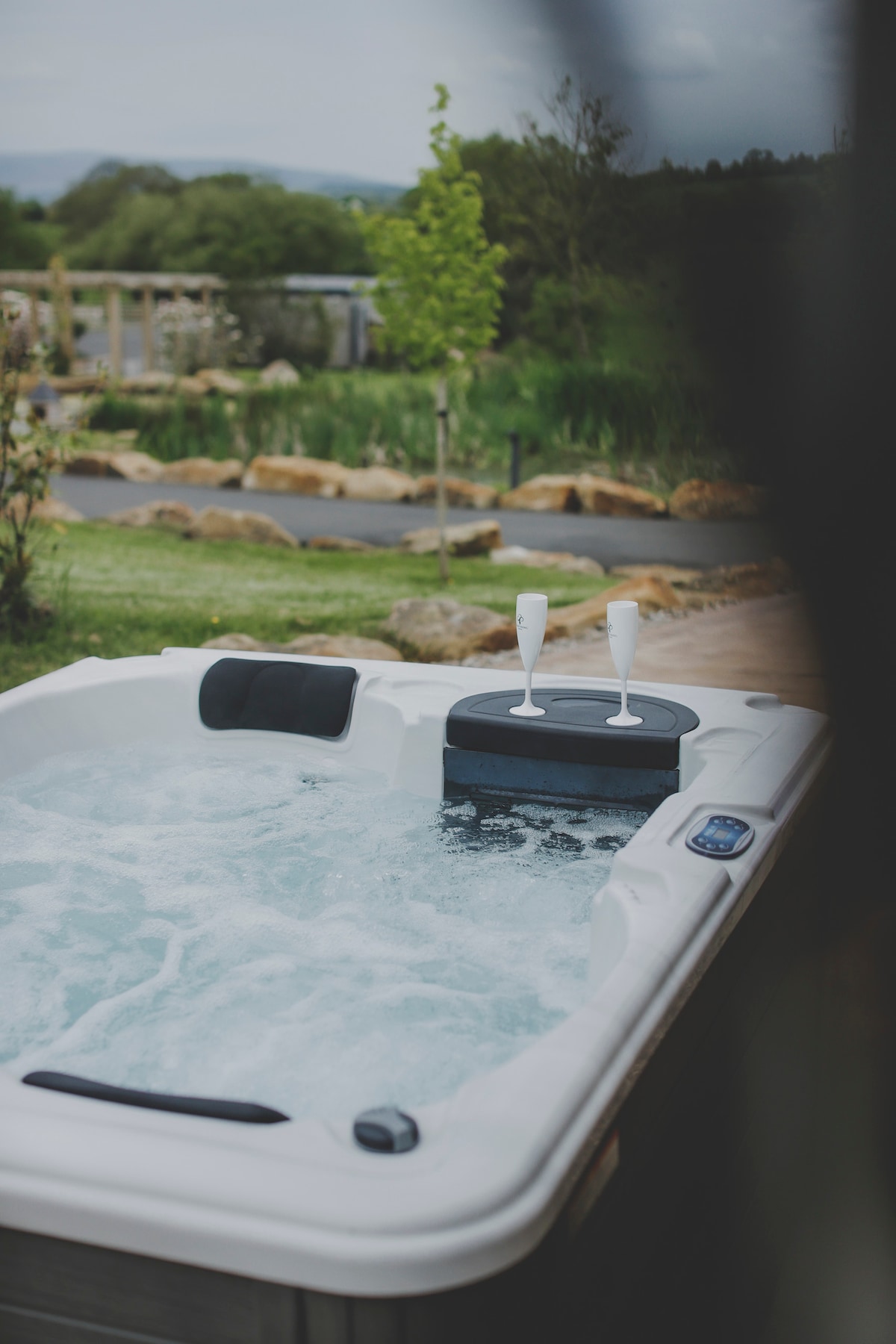 A hot tub is filled with bubbling water, featuring a small table in the center holding two champagne flutes. Surrounding greenery and a pebbled pathway enhance the peaceful outdoor setting.