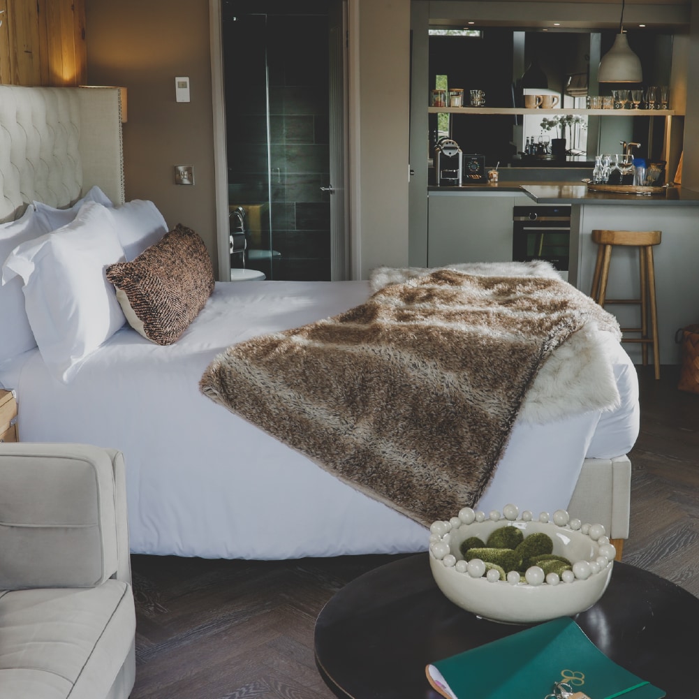 A spacious bed is adorned with a soft fur throw, accompanied by a decorative pillow. A cozy seating area features a light-colored armchair, with a dark coffee table displaying a bowl of decorative spheres. The kitchenette area includes wooden bar stools.