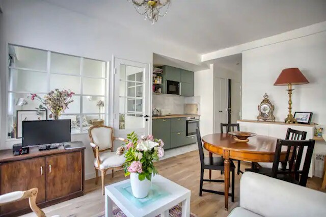The living area features a blend of modern and vintage furnishings, including a wooden table with chairs, a cozy sofa, and a vintage cabinet with a television. Natural light fills the space, highlighting the floral arrangement on the coffee table and a glimpse of the kitchen area.