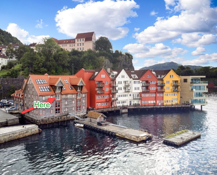 A waterfront view showcases a collection of colorful buildings, featuring shades of red, yellow, and white. The architecture reflects a coastal charm, with some structures displaying large windows. The serene water and surrounding greenery enhance the picturesque setting.