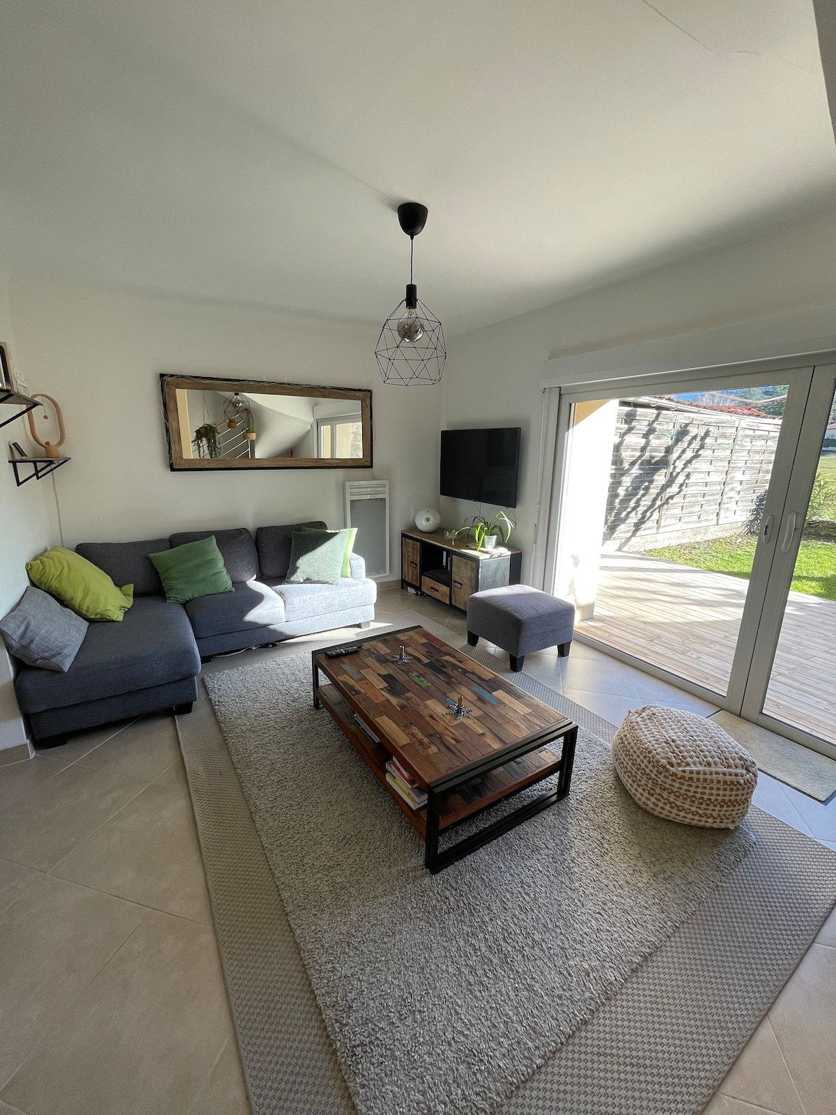 A light-filled living area features a grey sofa adorned with green cushions, complemented by a wooden coffee table. A large mirror hangs above the sofa, and a wall-mounted TV is positioned nearby. Glass doors open to a sunlit outdoor space, enhancing the room's spaciousness.