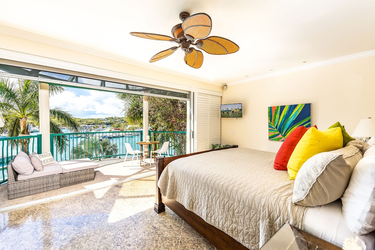 A spacious bedroom features a large bed with a neutral quilt. Colorful accent pillows brighten the bed, and large sliding doors open to reveal a private patio with views of the water. Sunlight illuminates the room, enhancing the serene atmosphere.