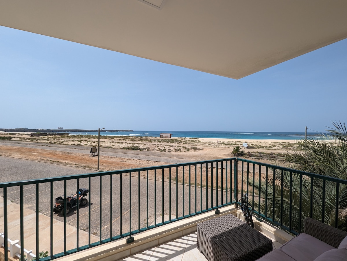 A spacious balcony offers views of distant sandy beaches and the ocean. The railing is green, complementing the outdoor furniture. Sunlight reflects off the water, while natural vegetation surrounds the area. The road below is visible, enhancing the sense of proximity to the beach.