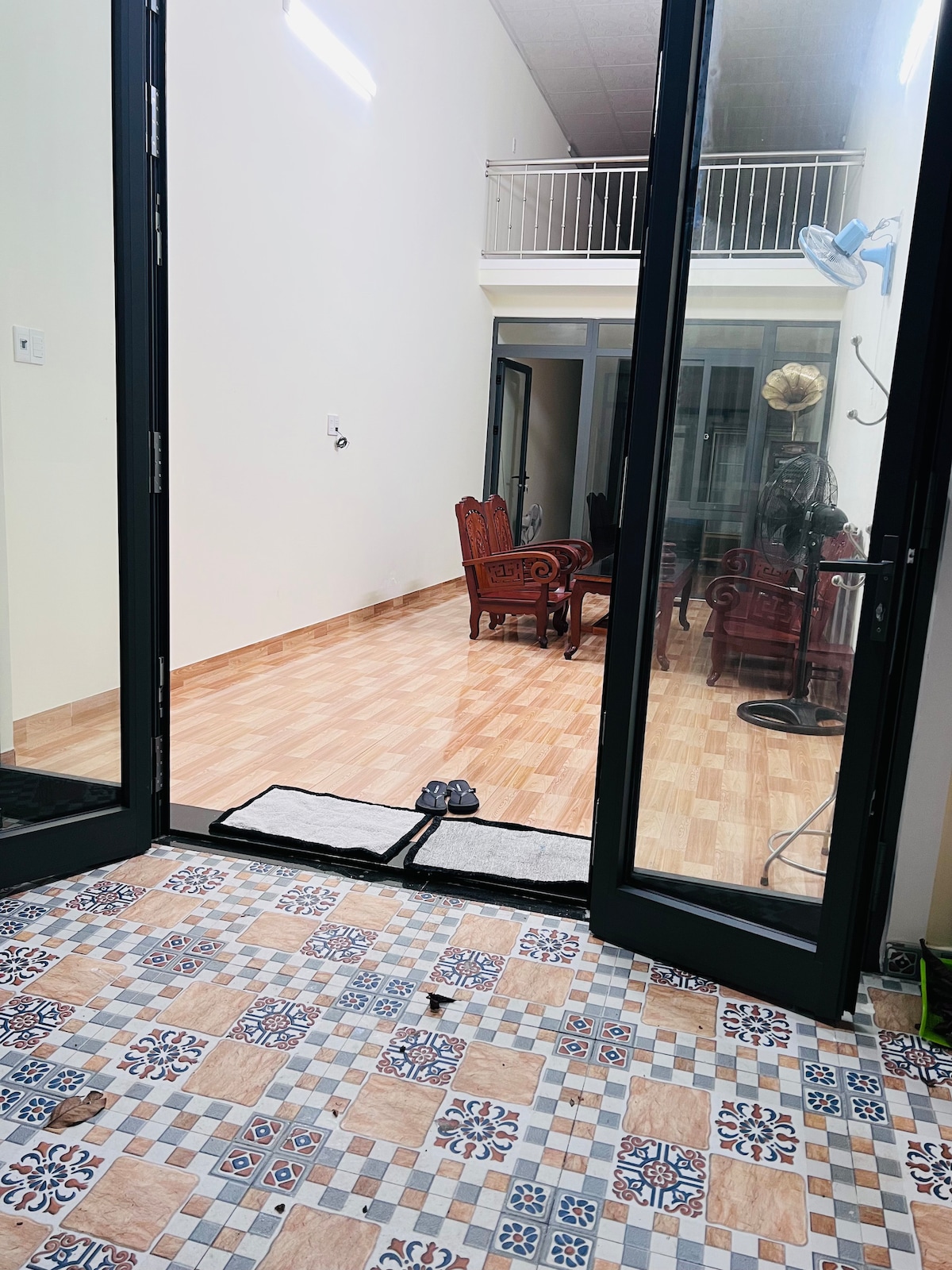 An open doorway provides a view into a spacious and well-lit living area. The floor features a combination of patterned tiles and wooden planks. Inside, a seating arrangement with dark wooden chairs can be seen, complemented by a large window allowing natural light to enter.