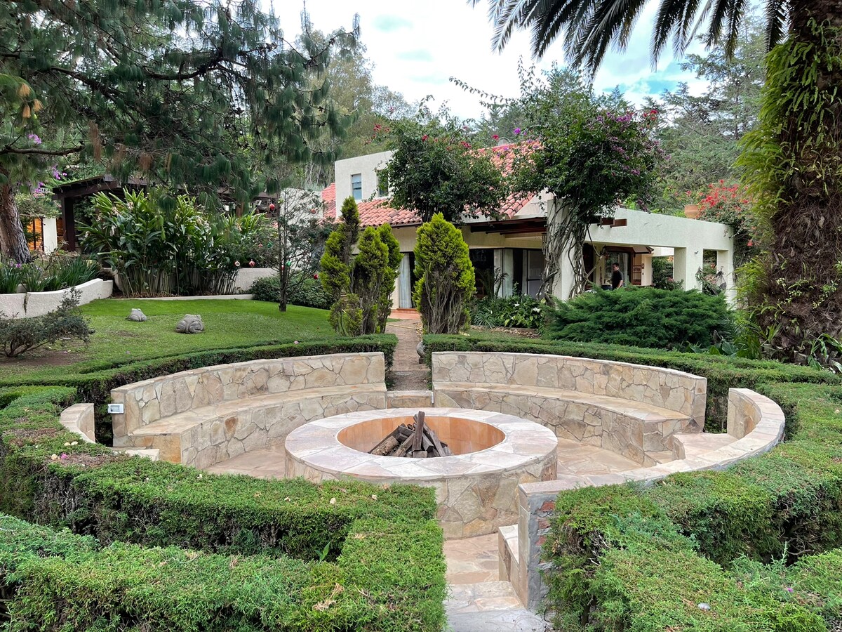 A circular stone fire pit is surrounded by neatly trimmed hedges and vibrant greenery. The landscaped area features various plants and trees, while a charming villa is visible in the background, blending with the serene outdoor environment.