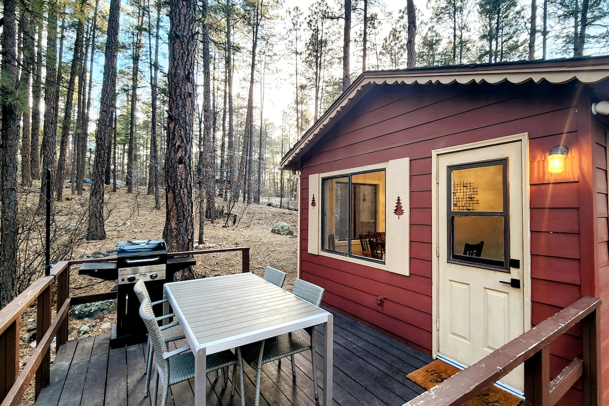 An outdoor deck features a gas grill and a dining table with six chairs, ideal for gatherings. The cabin's red exterior complements the natural surroundings of tall pine trees, creating a serene and inviting space for outdoor dining or relaxation.