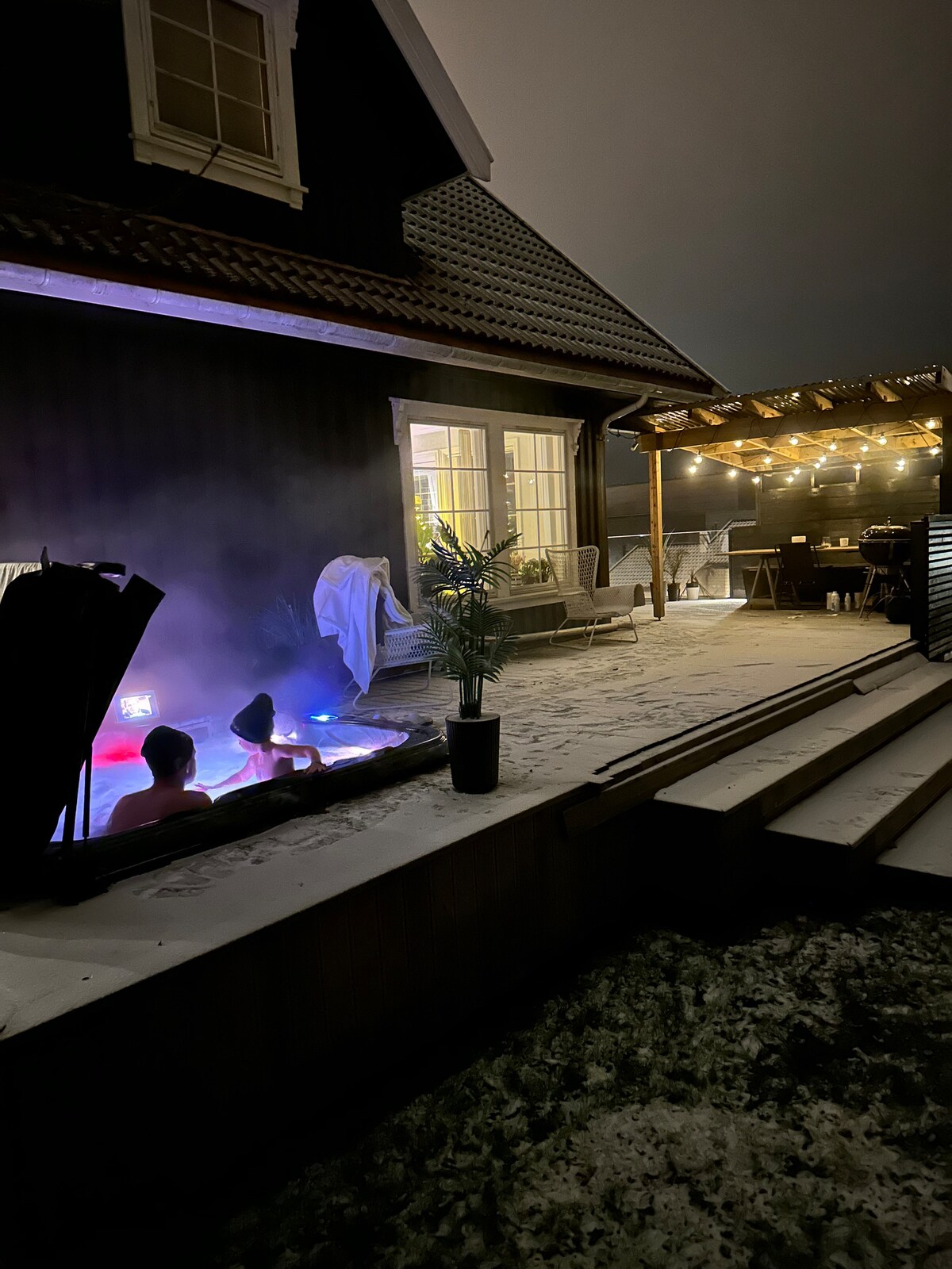 A cozy outdoor space is illuminated with warm lights from an adjacent patio area. A hot tub is visible with colorful lighting, surrounded by lightly falling snow. The house features large windows that provide a glimpse of indoor comfort and a tranquil atmosphere.