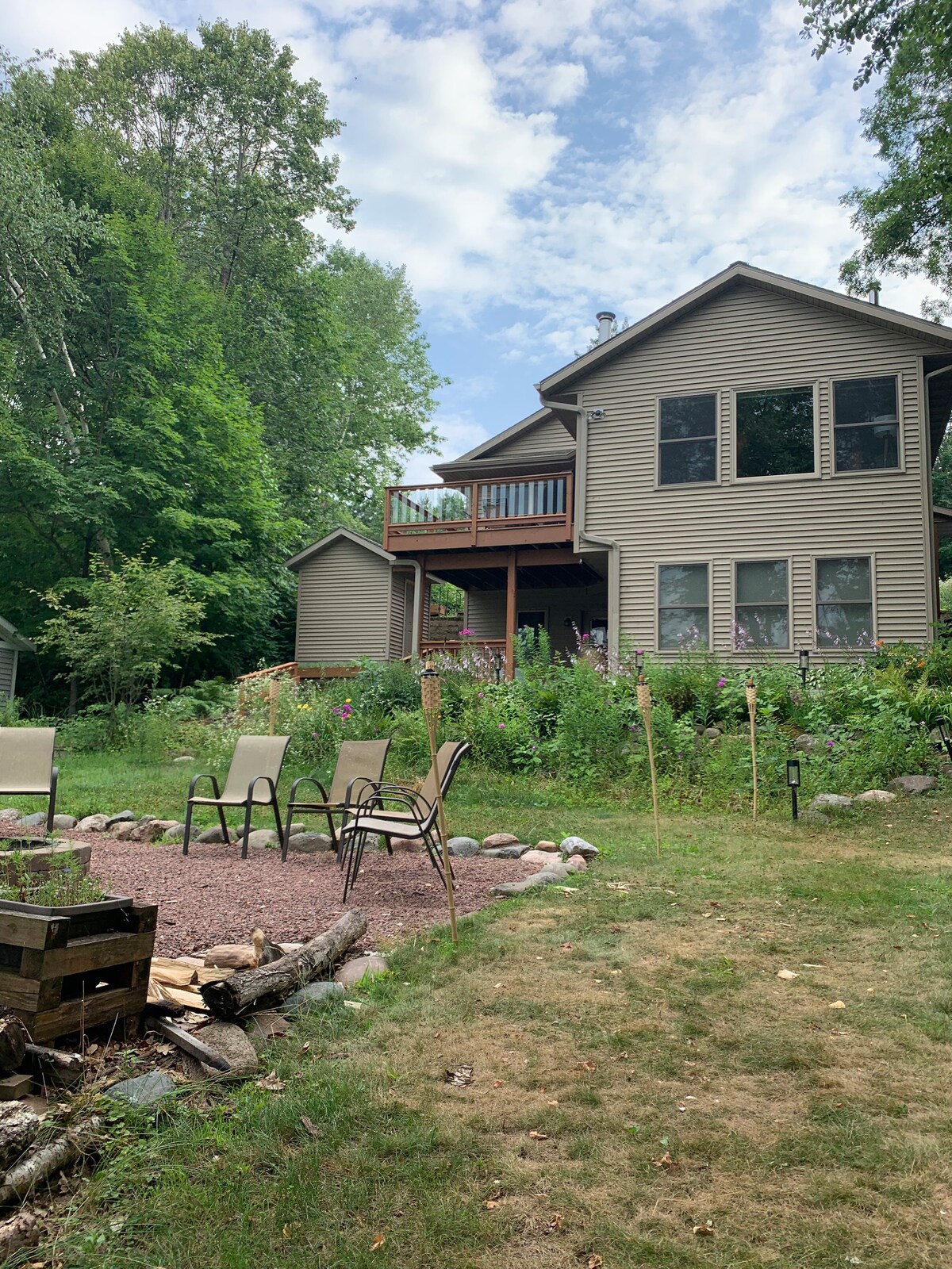 The rear view of the home is framed by lush greenery, showcasing a spacious deck above, accessible from the upper level. A gravel area features four chairs arranged around a fire pit, providing a cozy gathering space amidst the natural surroundings.