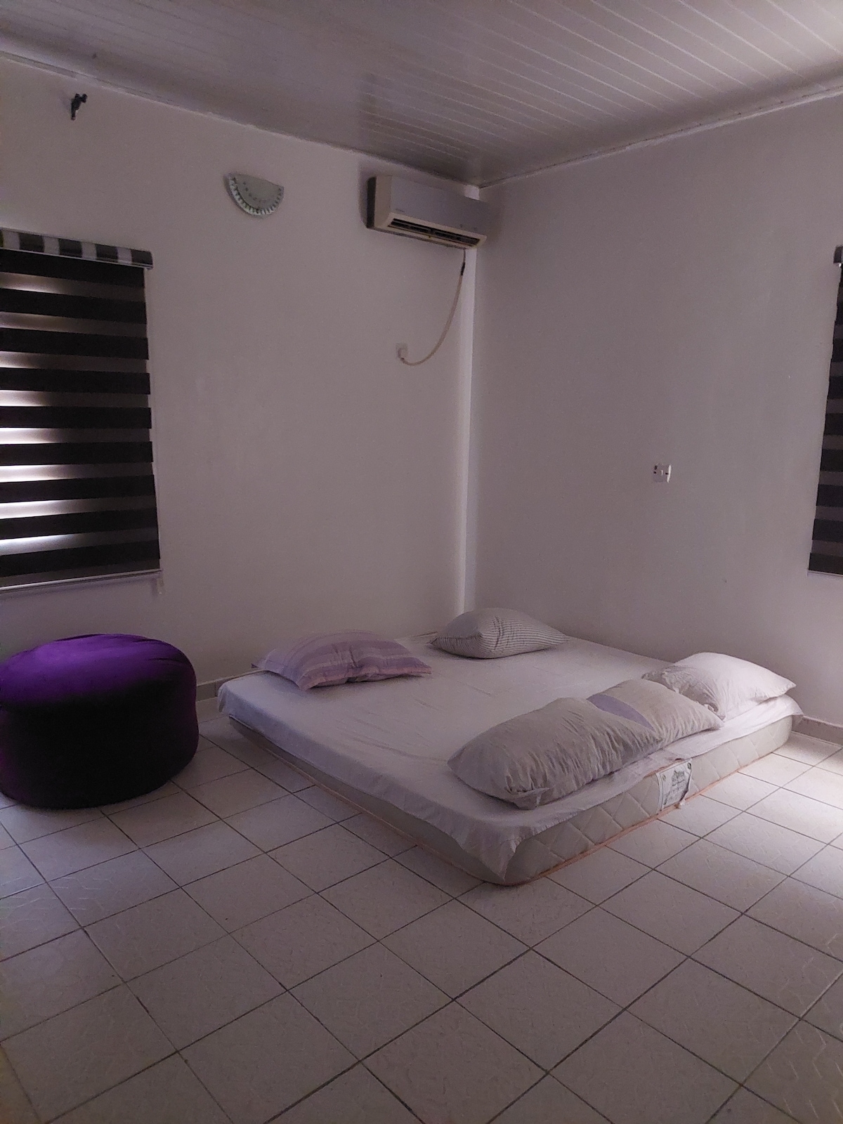 A minimalist sleeping area features a low mattress adorned with several soft pillows. A round purple ottoman is positioned nearby. Two windows dressed with dark striped curtains allow soft light to filter into the room.