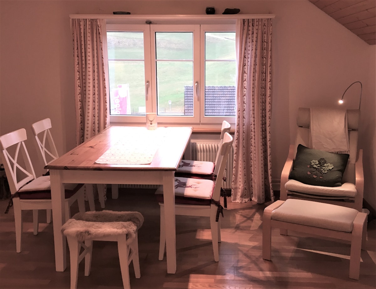 A dining area features a light wooden table surrounded by four white chairs and a small bench. A large window allows natural light to flood the space, framed by light-colored curtains. A comfortable armchair with a reading lamp is positioned nearby.