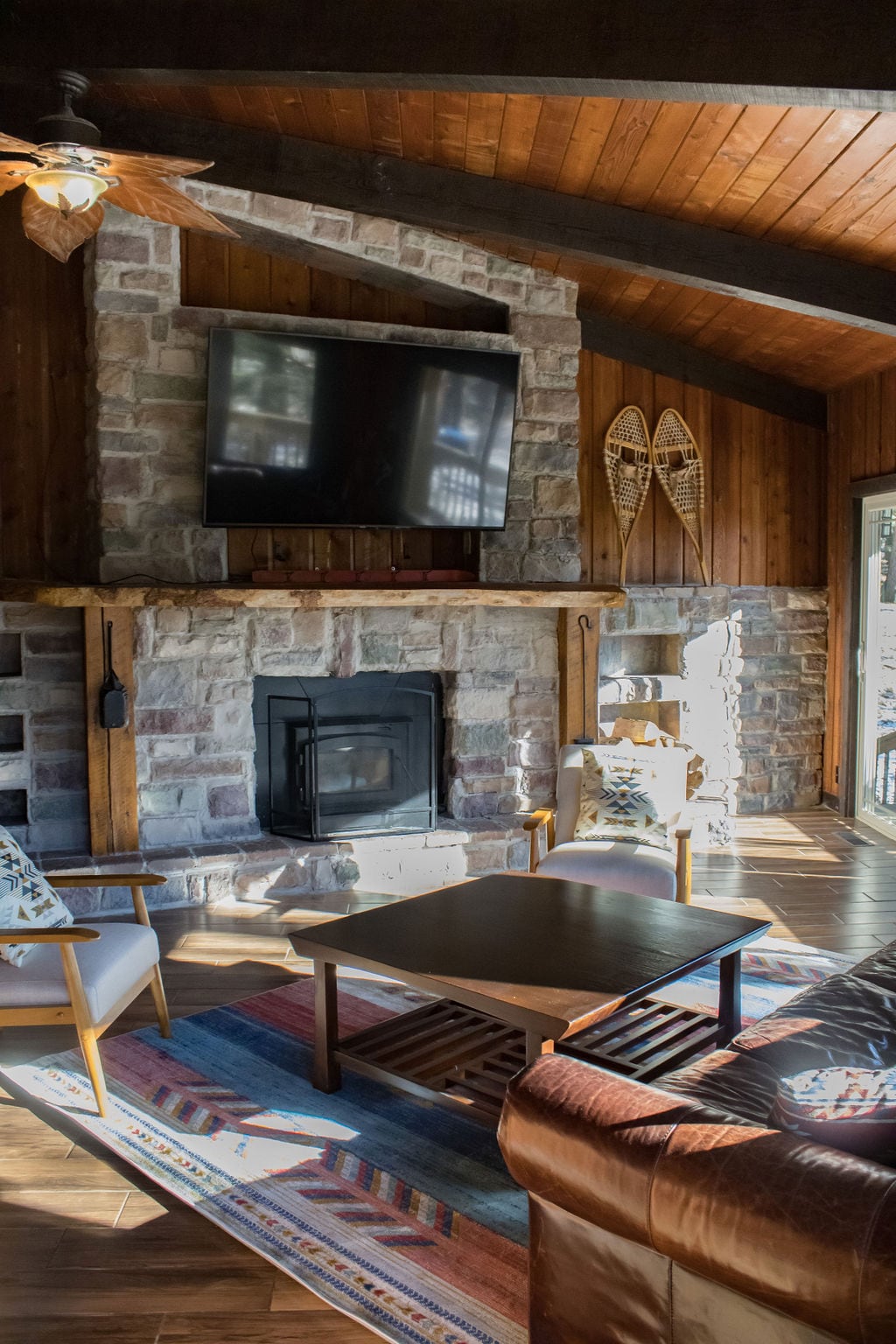 A cozy living space features a stone fireplace with a mounted television above it. A wooden ceiling complements the warm decor. Comfortable seating arrangements include a leather chair and patterned armchair. A coffee table sits atop a colorful rug, enhancing the inviting atmosphere.