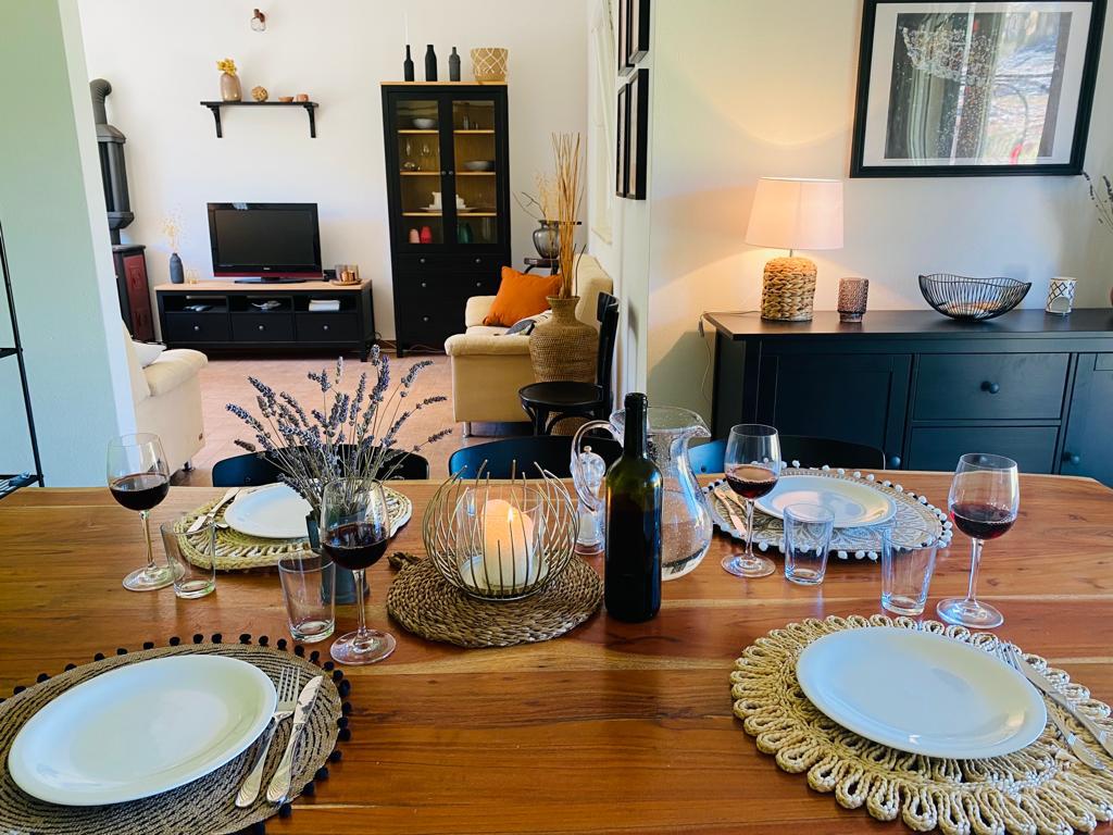 A dining table is set with plates and cutlery, accompanied by glasses of red wine. A centerpiece of dried flowers and a candle creates a subtle focal point. The open layout connects seamlessly to a cozy living area, featuring a television and comfortable seating.