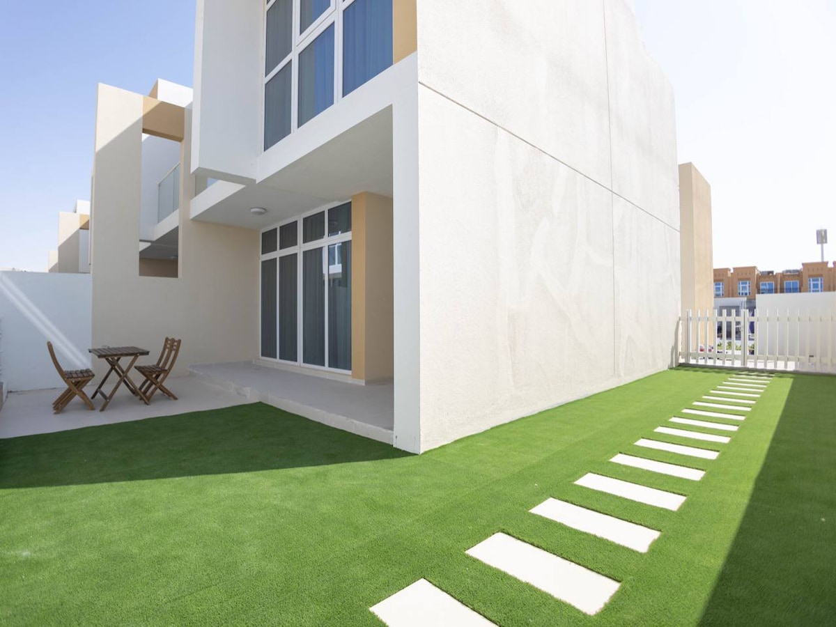 An outdoor area is shown featuring a well-maintained green lawn, complemented by a series of stepping stones leading toward a modern villa. A small wooden table with two chairs is positioned on a patio space, offering an inviting spot for relaxation.