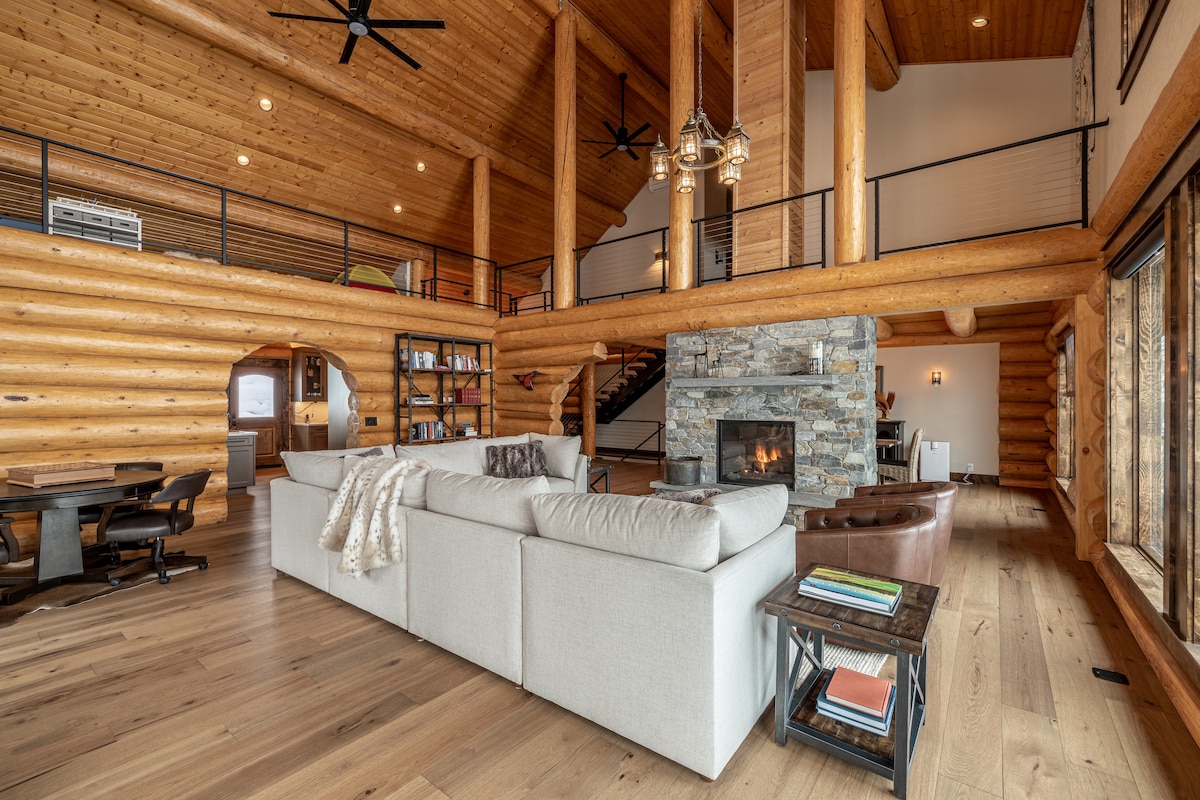An open living area is highlighted by high wooden ceilings and large windows, allowing natural light to fill the space. A stone fireplace serves as a central feature, with a cozy sectional sofa and a wooden coffee table positioned nearby. The warm wood tones enhance the inviting ambiance.