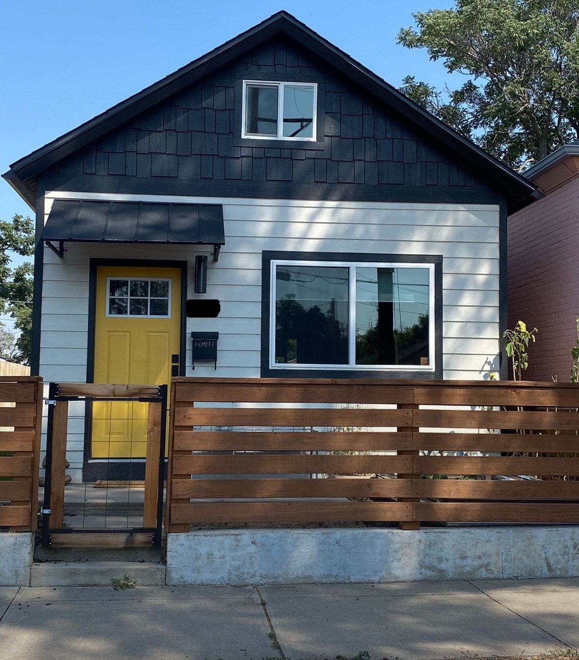 The exterior of a quaint house is displayed, featuring a black and white color scheme. A bright yellow door adds a pop of color, while a modern wooden fence offers privacy. Large windows provide natural light, and greenery is visible surrounding the entrance.