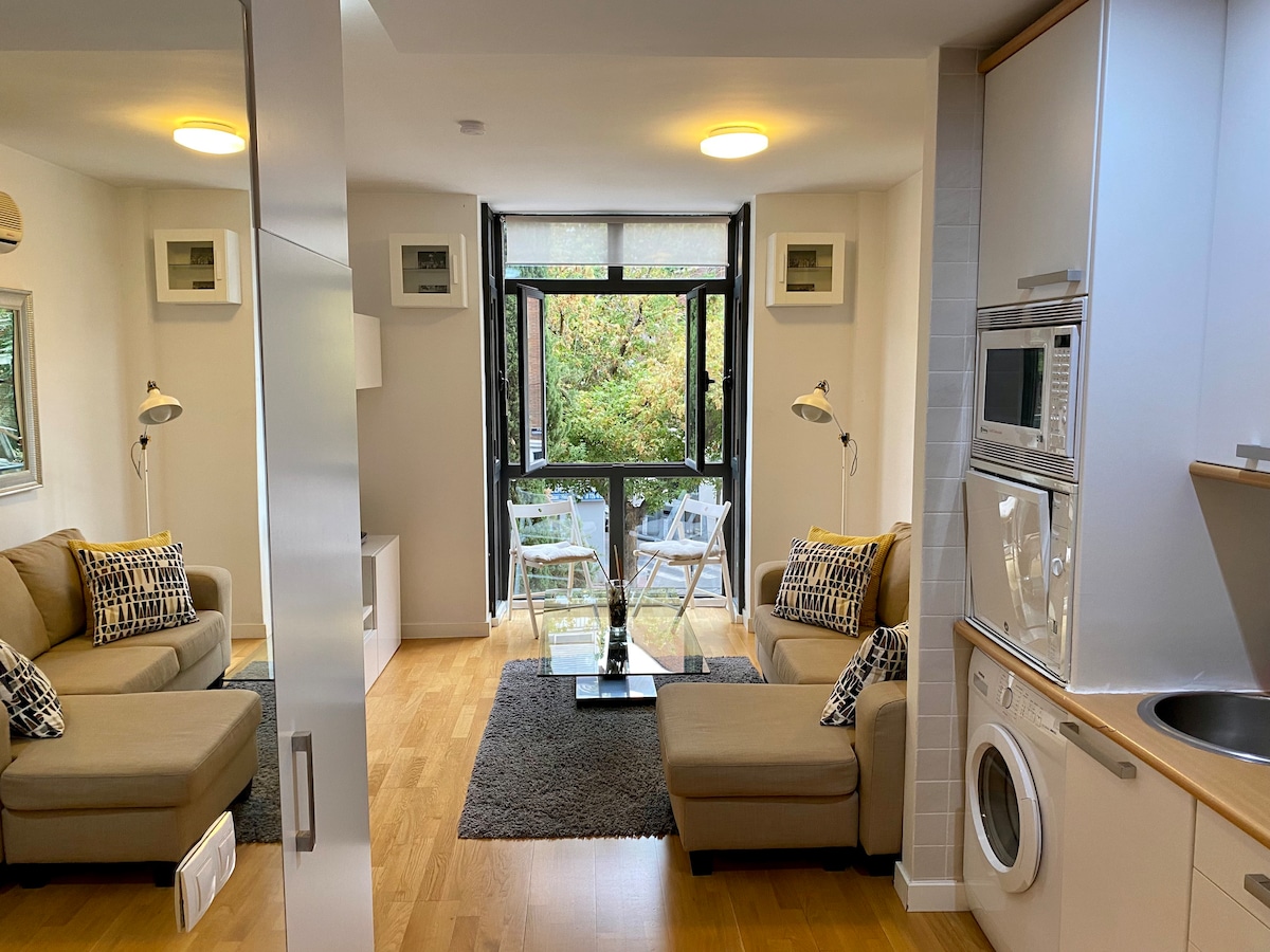 A modern living area is presented, featuring a comfortable sofa and a chaise longue. Large glass doors open to a balcony with two chairs and a small table, allowing natural light to flow in. An open kitchen area with essential appliances is visible to the side.