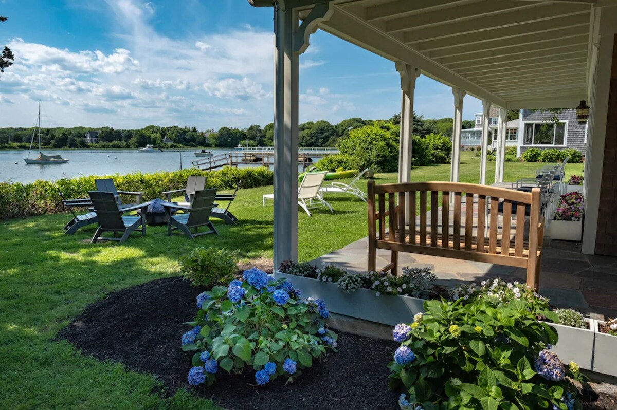 An inviting outdoor space features a wooden bench under a covered porch, surrounded by lush greenery and hydrangea flowers. Patterned lounge chairs are positioned on manicured grass, with a view of a calm waterway and sailboats in the distance, creating a serene environment.