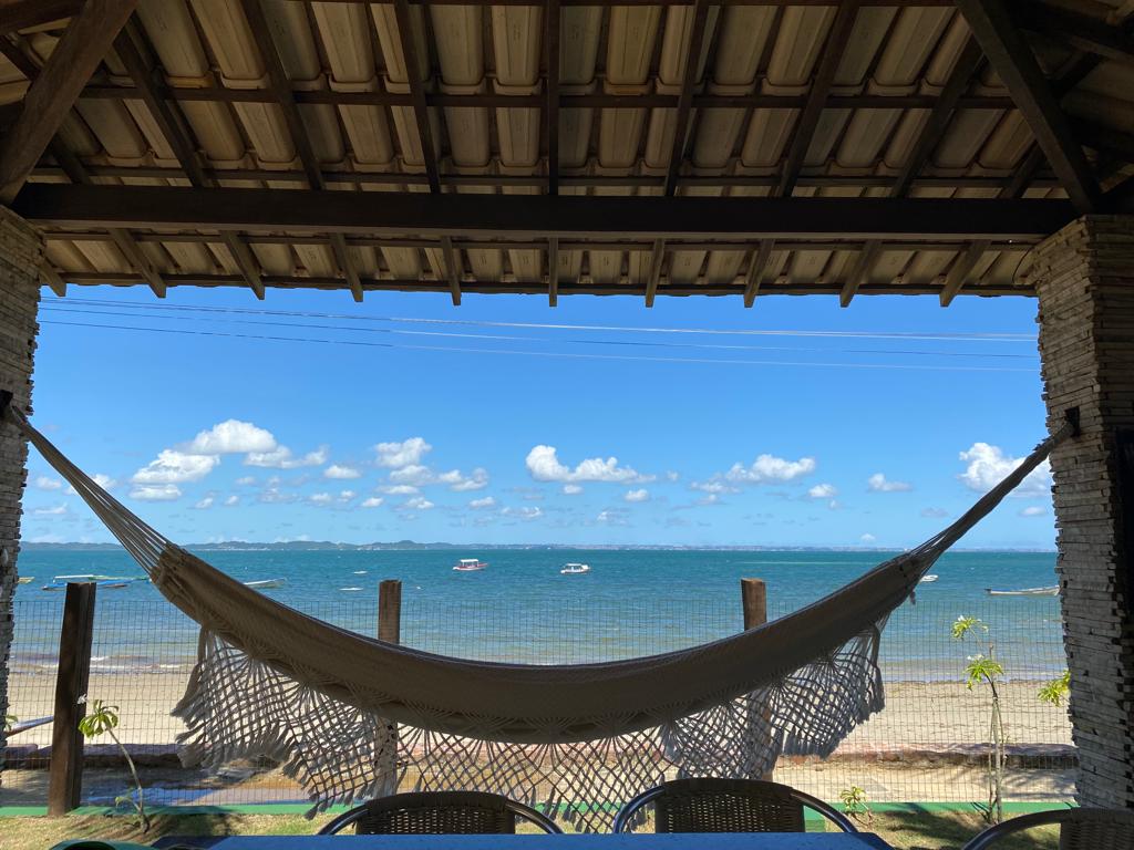 A spacious covered porch features a woven hammock suspended between two posts, offering a view of the serene beach and tranquil sea. Soft clouds drift across the blue sky, while boats are visible on the water. Two chairs are positioned nearby, inviting relaxation.