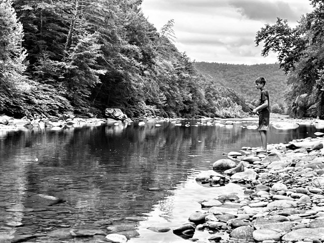 A child is seen standing at the water's edge of a gently flowing creek, surrounded by smooth stones. Lush greenery lines the banks, and distant mountains are visible under a cloudy sky. The image is presented in black and white, emphasizing the natural landscape.