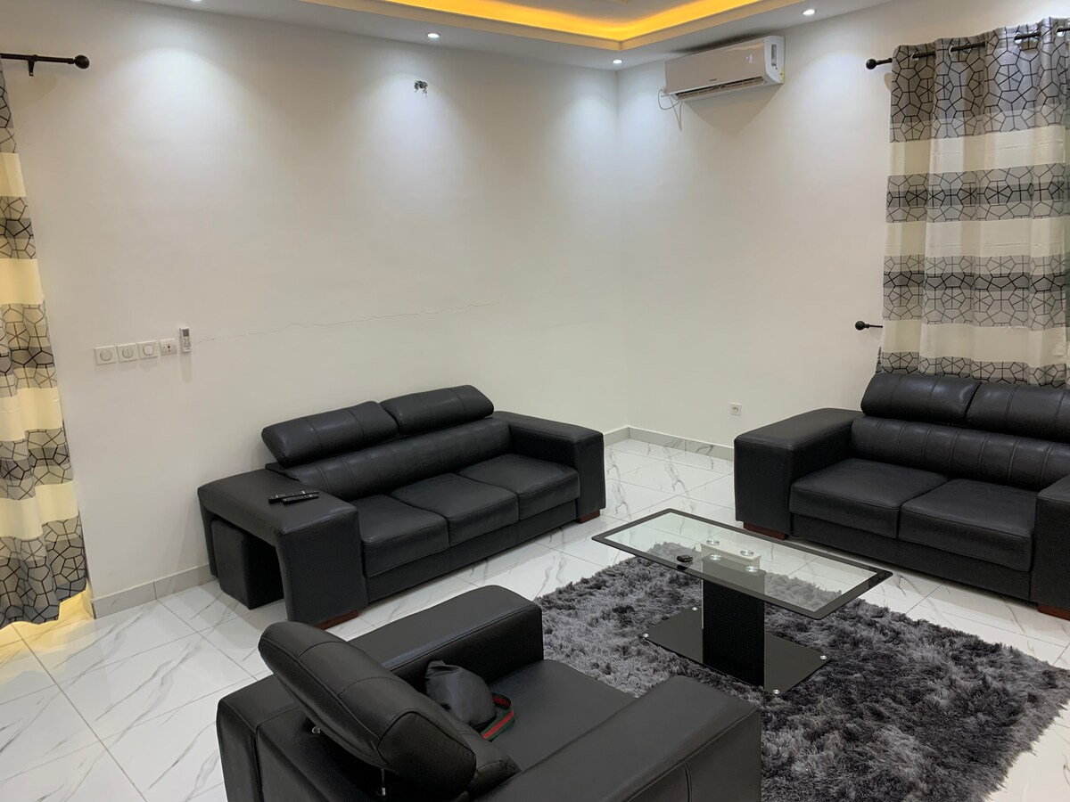 A modern living room features a sleek black leather sofa set arranged around a glass coffee table. The space is illuminated by soft lighting, complemented by dual-tone patterned curtains on either side of the windows. A plush grey rug sits beneath the coffee table.