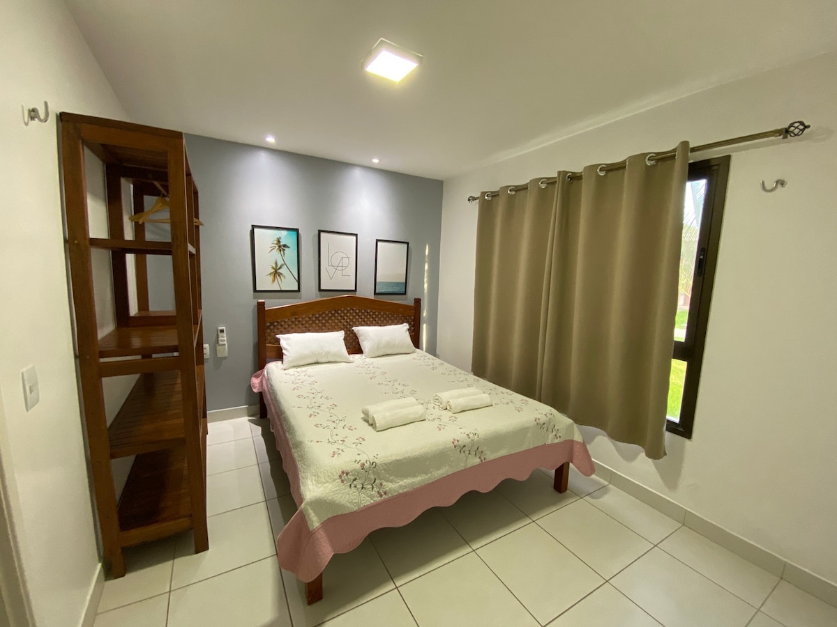 A well-lit bedroom features a spacious bed adorned with light linens and decorative pillows. A wooden shelving unit stands nearby, while natural light filters through a large window, framed by beige curtains. Minimalist wall art adds a touch of style to the gray accent wall.