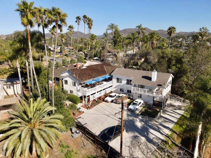An aerial view of the guest studio property reveals a spacious layout with palm trees surrounding the property. The large deck extends from the building, with a swimming pool visible in the backyard. Ample parking space is provided in front of the property.