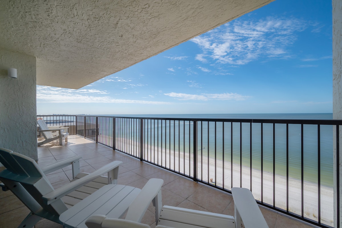 An expansive balcony showcases a stunning view of the Gulf of America, framed by a black railing. Two Adirondack chairs are positioned for relaxation, with a sandy beach visible below. The sky is clear, reflecting peaceful tones of blue and white.