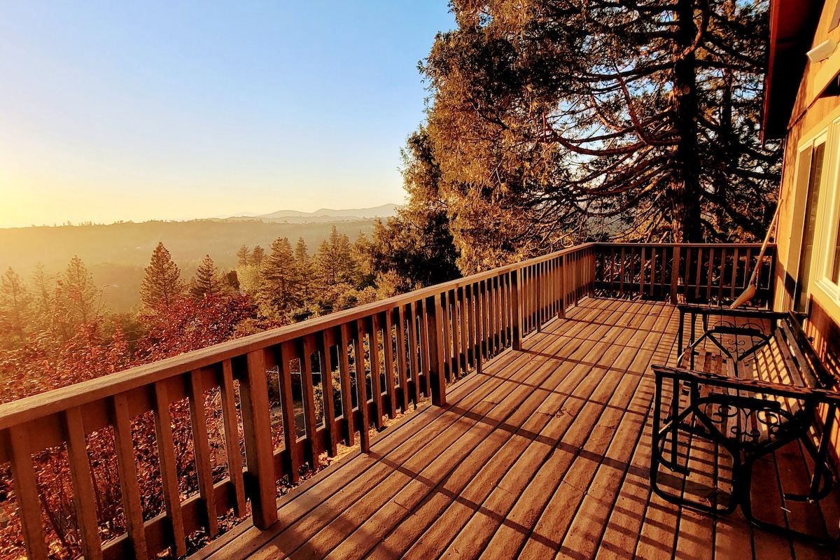 A spacious outdoor deck features wooden flooring and sturdy railings, providing a panoramic view of the valley and trees. Two chairs are positioned on the deck, inviting guests to relax and enjoy the tranquil setting during sunset.