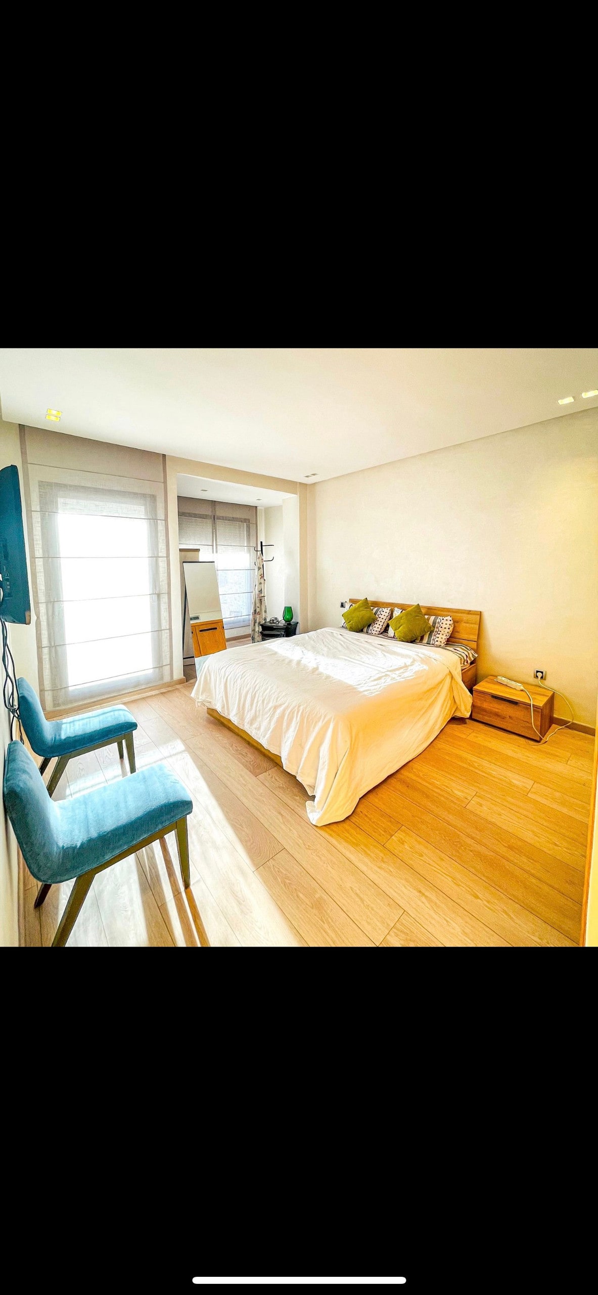 A bright sleeping area features a spacious bed with neutral bedding and accent pillows. Two blue chairs are positioned beside a wooden nightstand. Natural light fills the room through large windows covered with sheer curtains, highlighting the light wood flooring.
