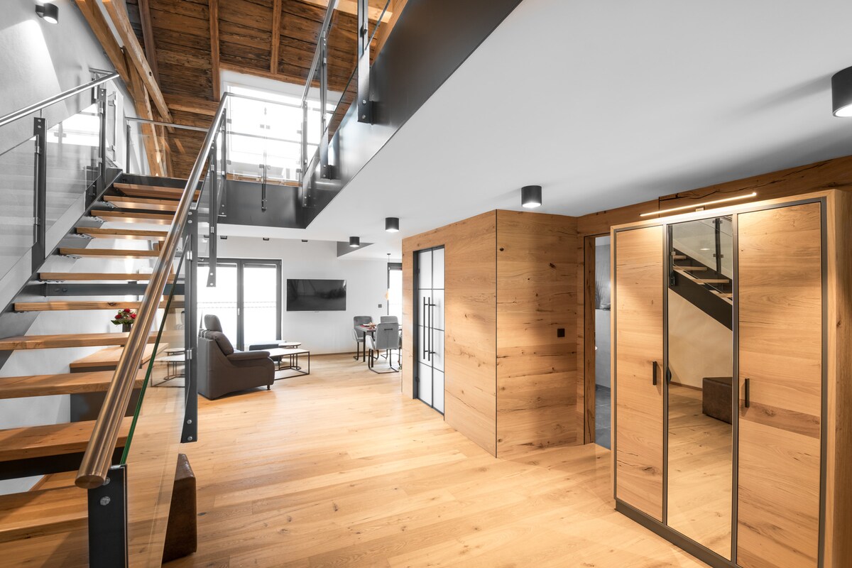 An airy open space is showcased with a wooden staircase leading to an upper level. Light-colored wooden flooring complements the contemporary decor, while large glass doors invite natural light. A modern seating area is visible in the background, alongside a welcoming dining setting.