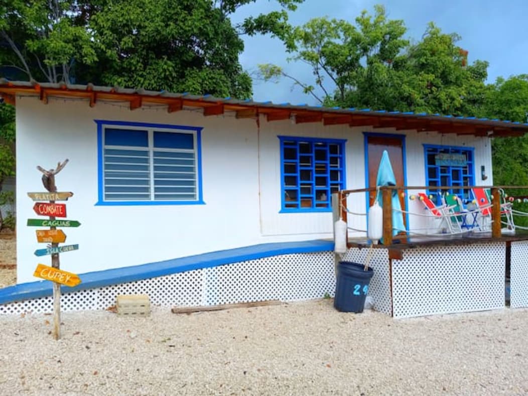 The charming exterior of the vacation home features a white façade accented with blue window frames. A wooden deck with colorful chairs overlooks the gravel yard. A directional signpost shows the way to local attractions, adding a welcoming touch to the entrance.