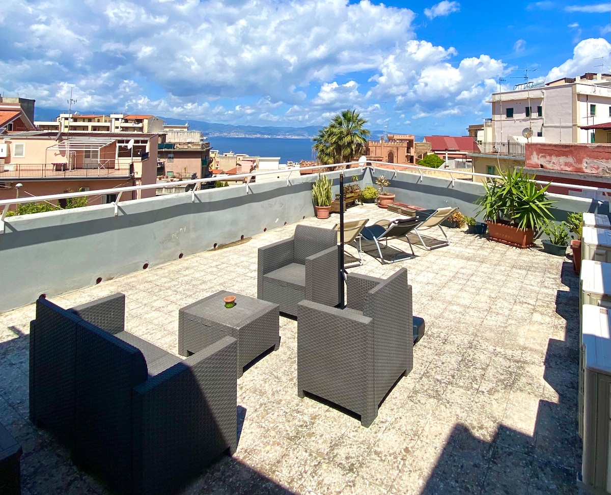 The spacious terrace is furnished with a comfortable seating area, including a table and chairs. Sun loungers are positioned for relaxation. A backdrop of the sea and palm trees is visible, accompanied by a bright sky scattered with clouds.