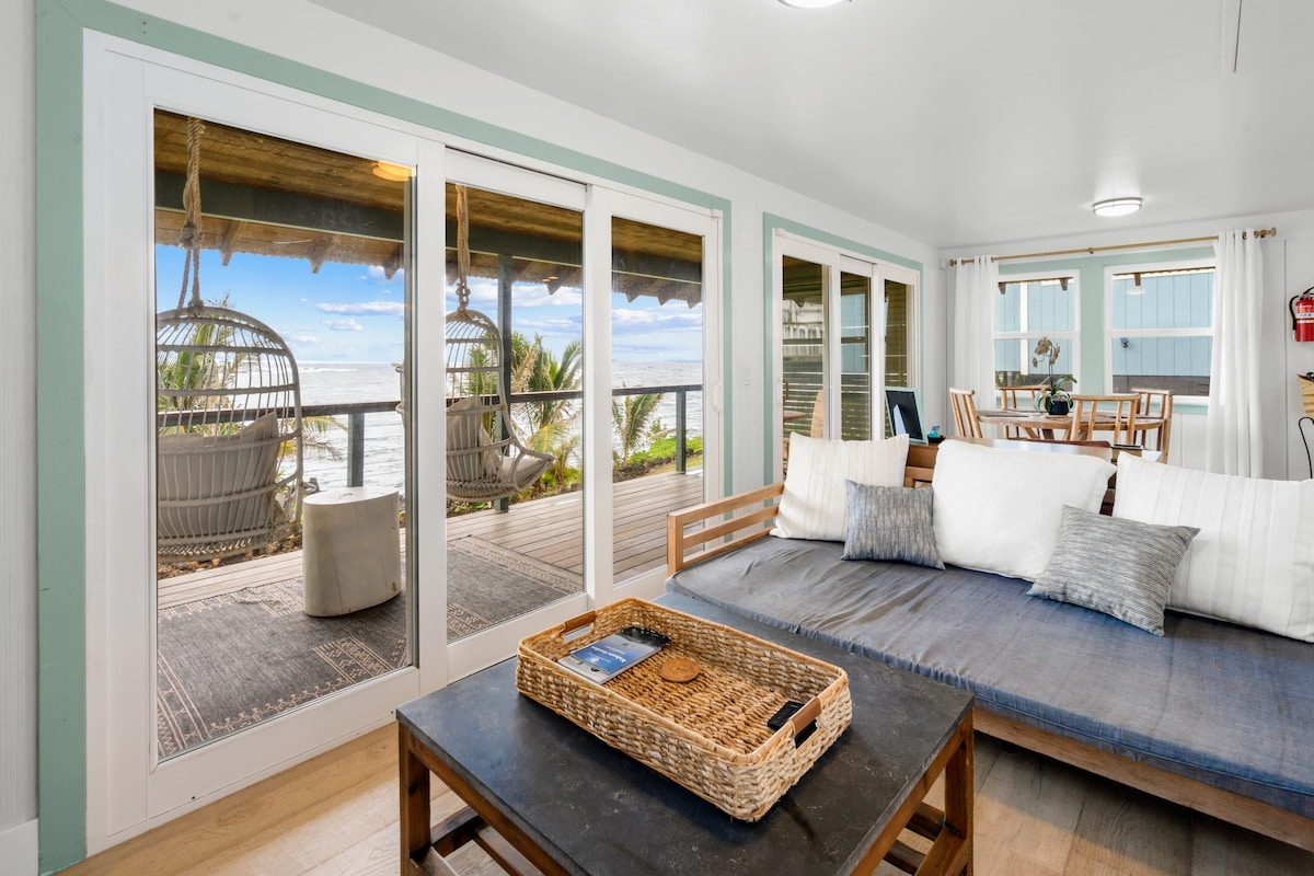 An open living space showcases a comfortable sofa and a coffee table with decorative items. Large glass doors provide a view of the covered ocean deck and lush palm trees outside, enhancing the connection to the outdoor environment.
