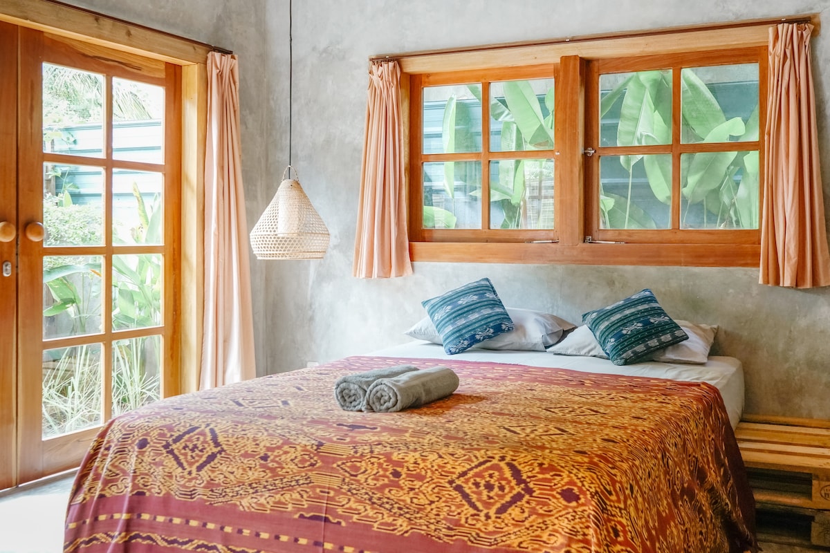 A bright bedroom features a large bed adorned with a colorful patterned blanket. Natural light pours in through two windows, and a woven pendant light hangs overhead. Towels are neatly rolled on the bed. A door leads to a green outdoor space.