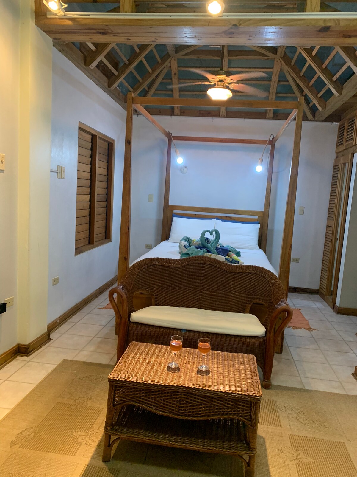 A spacious bedroom is framed by a wooden four-poster bed, complemented by a cozy seating area in a woven design. Soft lighting illuminates the room, while warm tones and tiled flooring create a comfortable ambiance. Complementary decor includes a decorative item on the bed.