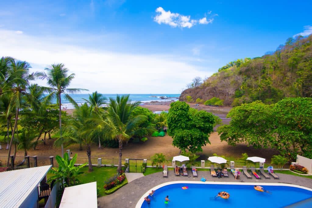An oceanfront view highlights the blue pool surrounded by tropical greenery. Palm trees and lush vegetation frame the scene, with the beach and ocean visible in the background. Clear skies and distant hills complete the serene coastal atmosphere.