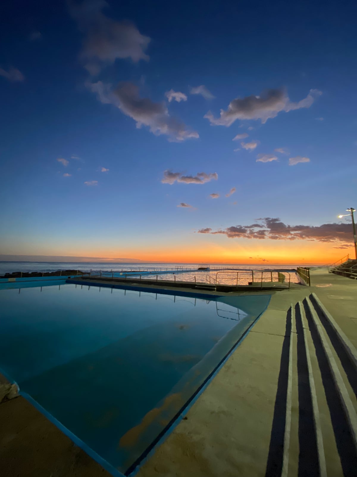 A tranquil swimming pool is visible at dusk, reflecting the vibrant colors of the sunset. The sky showcases soft clouds and gradients of orange and blue. The pool's edge is lined with steps leading down into the water, while the ocean is seen in the background.