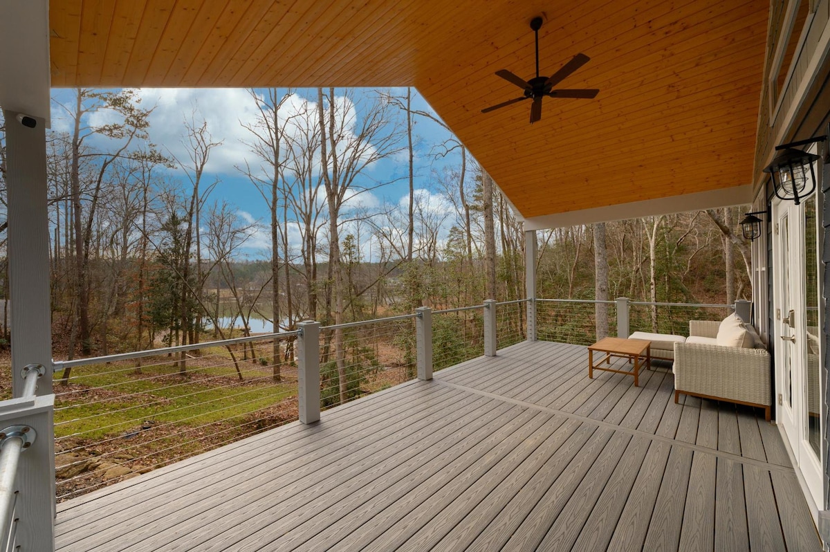 A spacious outdoor deck features a wooden floor and is partially shaded by a wooden ceiling. A comfortable seating area is arranged with a light-colored sofa and a coffee table. Surrounding trees provide a natural backdrop, with a glimpse of the lake visible in the distance.