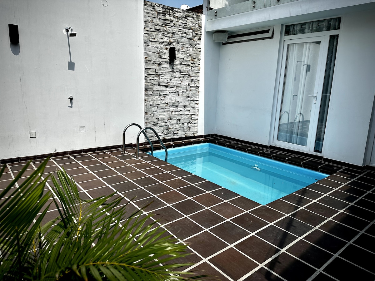 A private swimming pool is situated against a backdrop of textured stone walls. The pool's clear blue water is inviting, surrounded by dark tiled flooring. A glass door leads to an internal space, with greenery visible in the foreground.