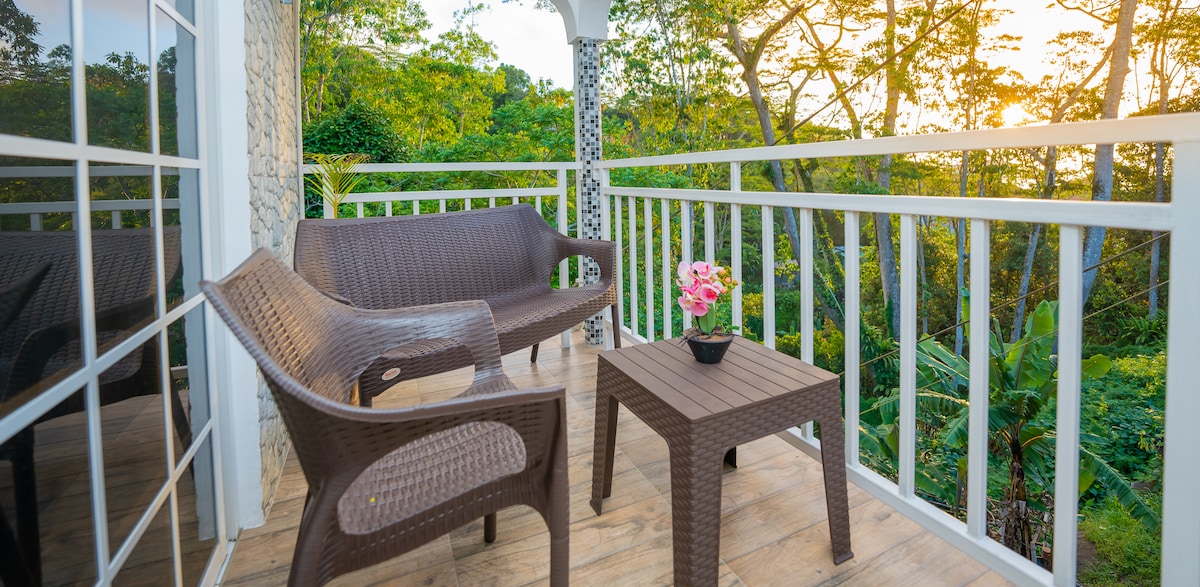 A private balcony features two lightweight, woven chairs and a small table, complemented by a potted pink flower. Natural light illuminates the space, with lush greenery visible in the background, creating a peaceful setting for relaxation.