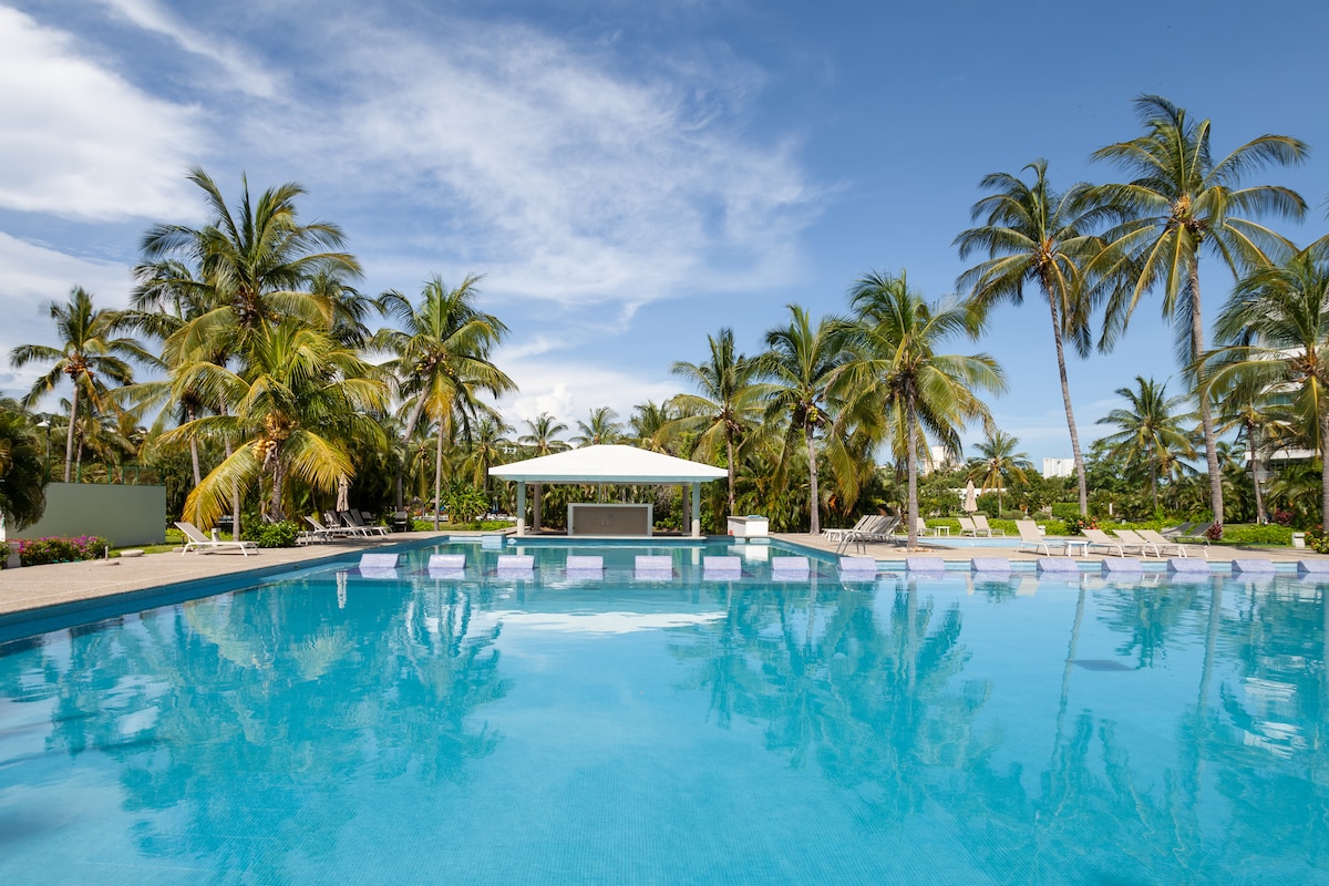A large swimming pool is surrounded by tropical palm trees, creating a relaxing atmosphere. A shaded structure is positioned near the poolside, offering relief from the sun. Light reflects off the water, enhancing the tranquil environment, suitable for leisure and family enjoyment.
