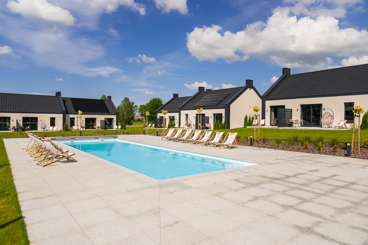 A spacious outdoor area features a long, inviting swimming pool surrounded by well-maintained greenery. Sun loungers are neatly arranged along the poolside, offering a place to relax. Modern accommodations with large windows can be seen in the background under a clear blue sky.